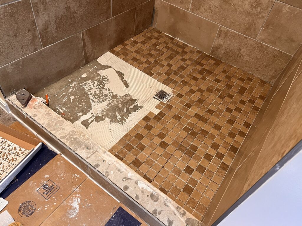Mosaic tile shower floor installation in progress with thinset mortar being applied around drain during bathroom remodel